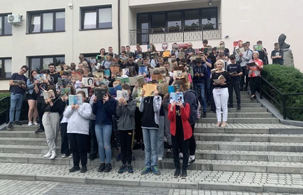 Fotografia barwna: grupa uczniów i nauczycieli biorących udział w akcji „Jak nie czytam, jak czytam”. Zdjęcie wykonano na tle budynku głównego szkoły. 