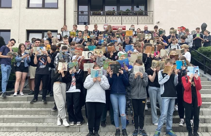 Fotografia barwna: grupa uczniów i nauczycieli biorących udział w akcji „Jak nie czytam, jak czytam”. Zdjęcie wykonano na tle budynku głównego szkoły. 