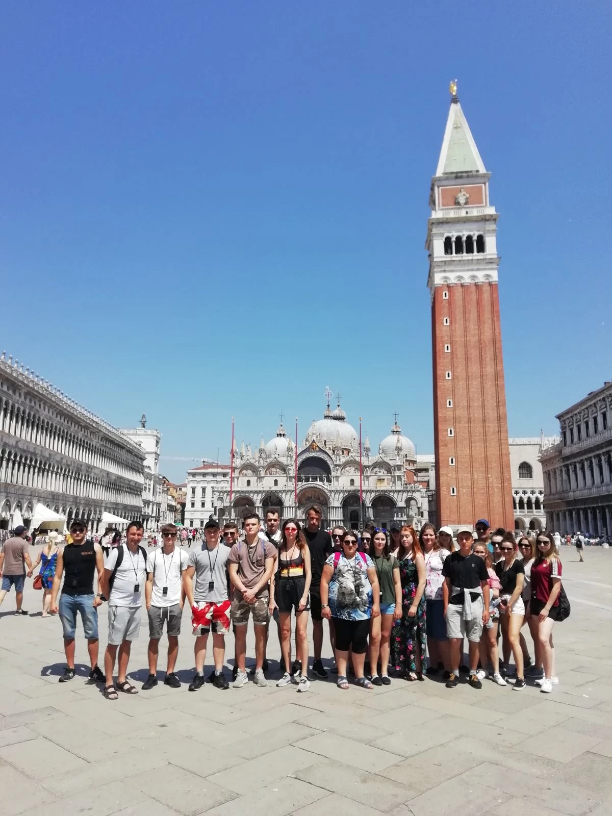 grupa ludzi pozująca do zdjęcia przed wysoką wieżą z Piazza San Marco w tle