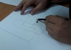 Zbliżenie na kartkę i dłonie szkicującego przestrzenne figury geometryczne.