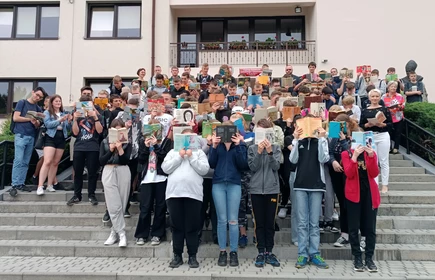 Fotografia barwna: grupa uczniów i nauczycieli biorących udział w akcji „Jak nie czytam, jak czytam”. Zdjęcie wykonano na tle budynku głównego szkoły. 
